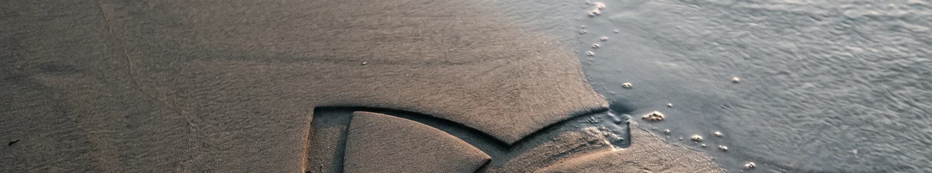 The Umber ribbon-knot mark carved into wet beach sand, the incoming tide washing in beside it.